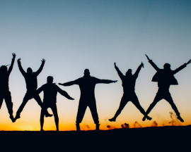 A group of silhouetted people jumping in celebration against a warm orange sunset.